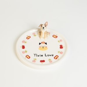 May include: A white ceramic plate with a raised edge, featuring a small dog figurine on top. The plate is decorated with red and pink floral and fruit designs, and the text "Ylvie Love".
