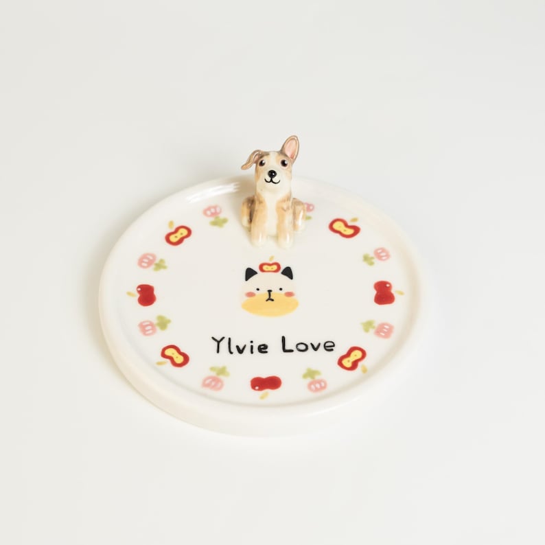 May include: A white ceramic plate with a raised edge, featuring a small dog figurine on top. The plate is decorated with red and pink floral and fruit designs, and the text "Ylvie Love".