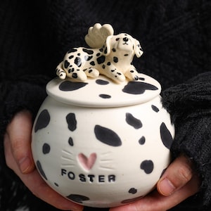 Op de afbeelding: Witte keramische pot met deksel, versierd met een Dalmatische hond figuur met zwarte vlekken. De pot heeft zwarte vlekken en het woord "FOSTER" onder een roze hart. De pot wordt in de handen van een persoon gehouden.