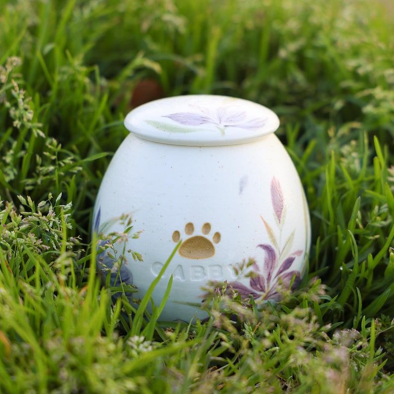 May include: White ceramic pet urn with a floral design and a paw print. The urn is inscribed with the name "Abby".