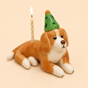 Könnte beinhalten: Eine Beagle-Figur aus Keramik in liegender Position, die einen grünen Partyhut mit dunklen Punkten trägt. Der Hund hat braun-weißes Fell, schwarze Augen und eine braune Nase. Eine dünne, brennende Geburtstagskerze steckt in einer Öffnung am Rücken.