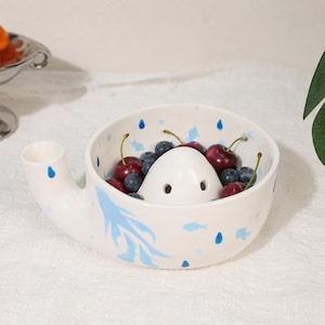 Könnte beinhalten: Eine weiße Keramikschale für Beeren mit seitlichem Ausguss und einem zentralen Kegel mit Abflusslöchern. Die Außen- und Innenseite sind mit hellblauen Fischen und dunkelblauen Regentropfen bemalt und mit frischen Kirschen und Blaubeeren gefüllt.