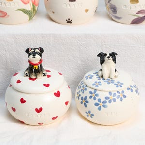 May include: Two ceramic jars with lids, one decorated with red hearts and a dog figurine, the other with blue flowers and a dog figurine. The jars are white with a glossy finish. The text "FOSTER" and "117" are visible on the jars.