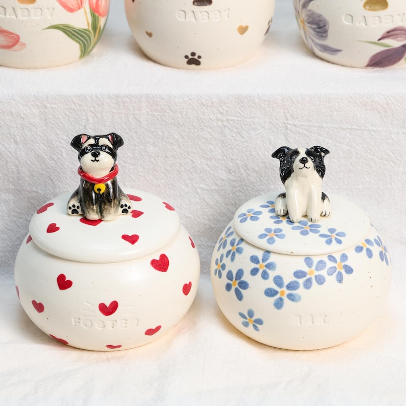 May include: Two ceramic jars with lids, one decorated with red hearts and a dog figurine, the other with blue flowers and a dog figurine. The jars are white with a glossy finish. The text "FOSTER" and "117" are visible on the jars.