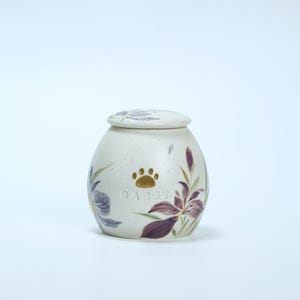 May include: White ceramic pet urn with a floral design featuring purple and green flowers. The urn has a gold paw print and the name "Abby" engraved on the front.