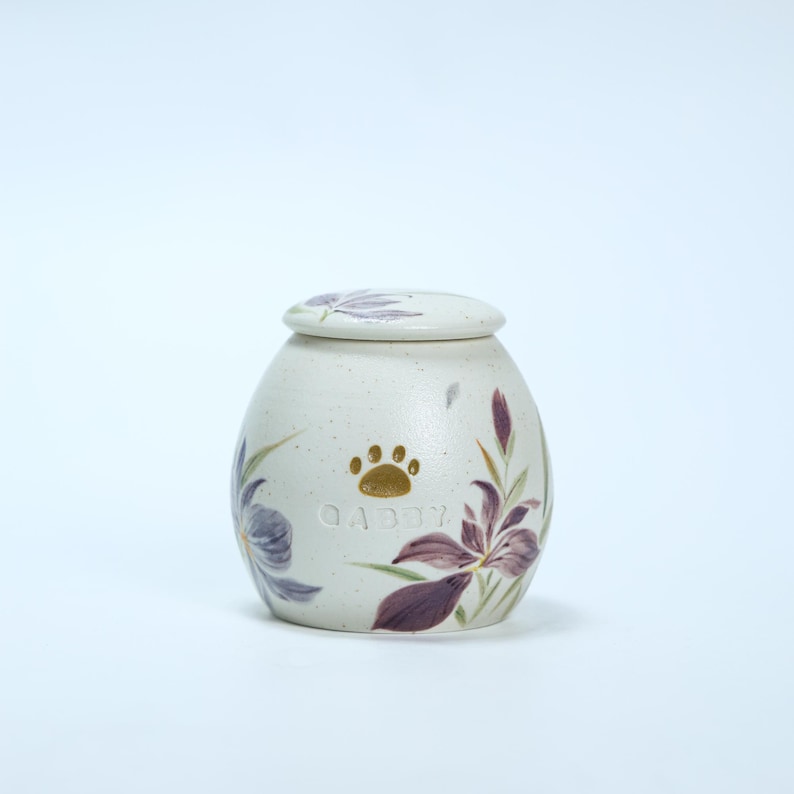 May include: White ceramic pet urn with a floral design featuring purple and green flowers. The urn has a gold paw print and the name "Abby" engraved on the front.