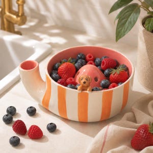 May include: A ceramic bowl with vertical orange and white stripes, featuring a pink interior and a small sculpted brown dog figure perched on the rim. The bowl has a side handle and contains a mix of fresh blueberries, raspberries, and strawberries.
