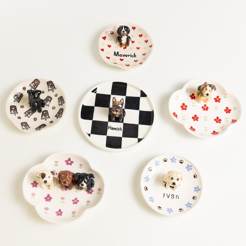 May include: A selection of decorative ceramic plates, each adorned with a dog figurine. The plates showcase diverse designs, such as floral, heart, and chequered patterns. Some plates are inscribed with names like "Maverick" and "Hamish".