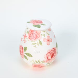 Op de afbeelding: Een witte, eivormige keramische urn met een deksel, versierd met roze rozen en groene bladeren. Het woord "Bella" staat op de urn. Het bloemmotief bedekt het gehele oppervlak en creëert een delicate en vrouwelijke uitstraling.