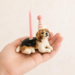 Könnte beinhalten: Eine Kerzenhalter-Figur aus Keramik in Form eines liegenden Beagles mit schwarzem, braunem und weißem Fell. Der Hund trägt einen kleinen, rosa-weiß gestreiften Partyhut auf dem Kopf und auf seinem Rücken steckt eine dünne, hellrosa Geburtstagskerze.