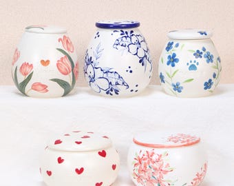 May include: Five white ceramic jars with lids, each adorned with unique floral designs. The jars feature hand-painted tulips, blue flowers, paw prints, red hearts, and pink floral patterns. The jars vary in size and shape.