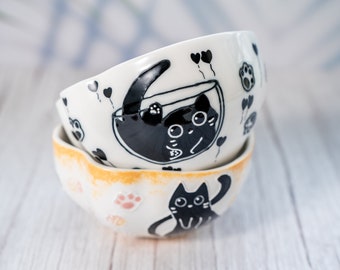 May include: Two white ceramic bowls with black cat designs. The top bowl features a black cat with a balloon and the bottom bowl features a black cat with paw prints.