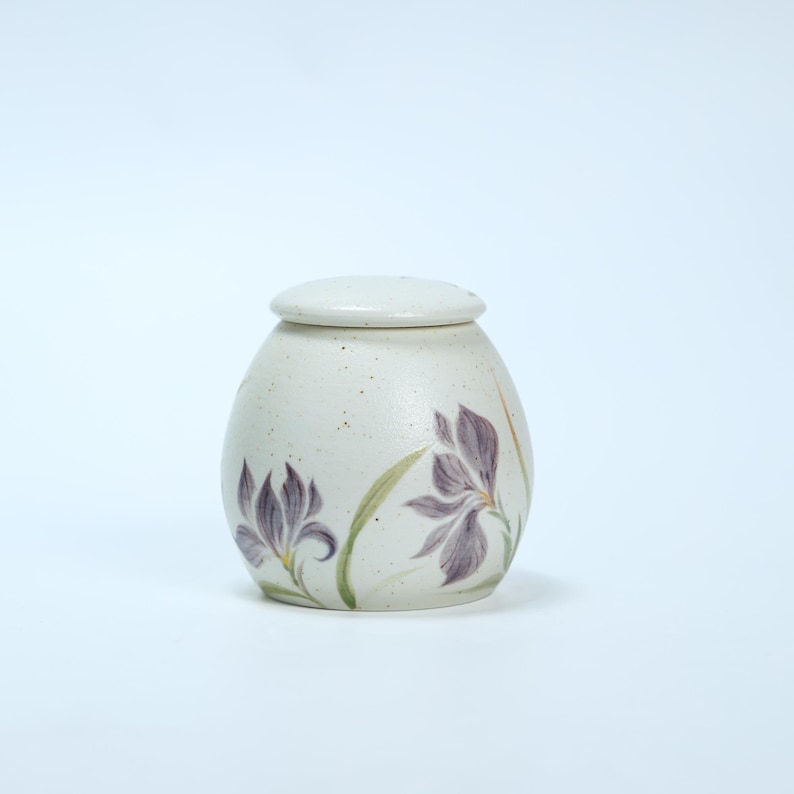 May include: A white ceramic jar with a lid, decorated with purple flowers and green leaves. The jar has a speckled finish.