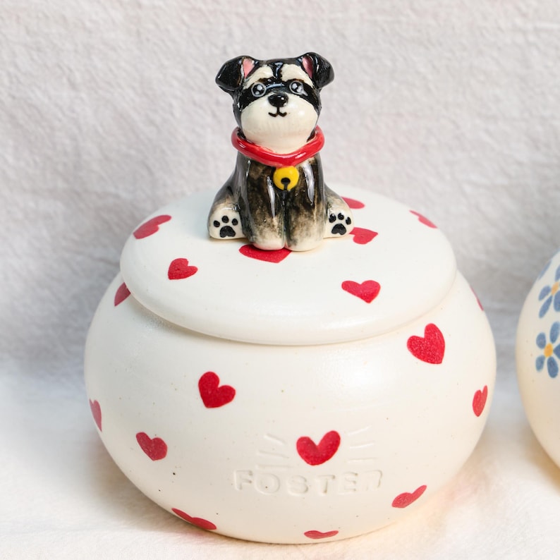 May include: A white ceramic jar with a lid, adorned with red hearts. A small, hand-painted dog figurine sits on the lid. The dog has black and grey fur, a red collar, and a yellow bell. The word "FOSTER" is printed on the jar.