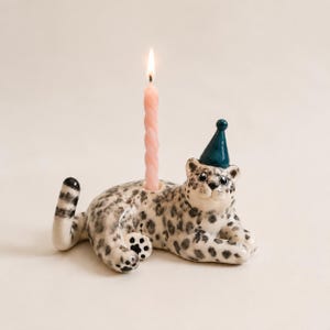 May include: A ceramic leopard figurine candle holder with a white body and black spots, wearing a small teal party hat. The leopard is in a reclining position with its tail raised. A single, thin, twisted light pink candle is lit in the holder.