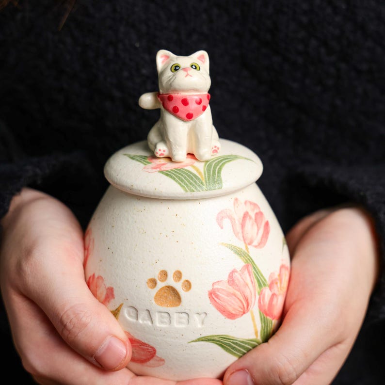 May include: A cream ceramic pet urn with a lid, decorated with pink tulip and green leaf floral patterns. A white cat figurine with a red polka-dot bandana sits on the lid. The urn is inscribed with the name "Gabby" and a paw print.
