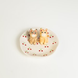 May include: A small, off-white ceramic dish with a scalloped edge, decorated with red cherries and green leaves. Two small cat figurines, one tan and one orange, sit on the dish. The cats have white bows on their heads.