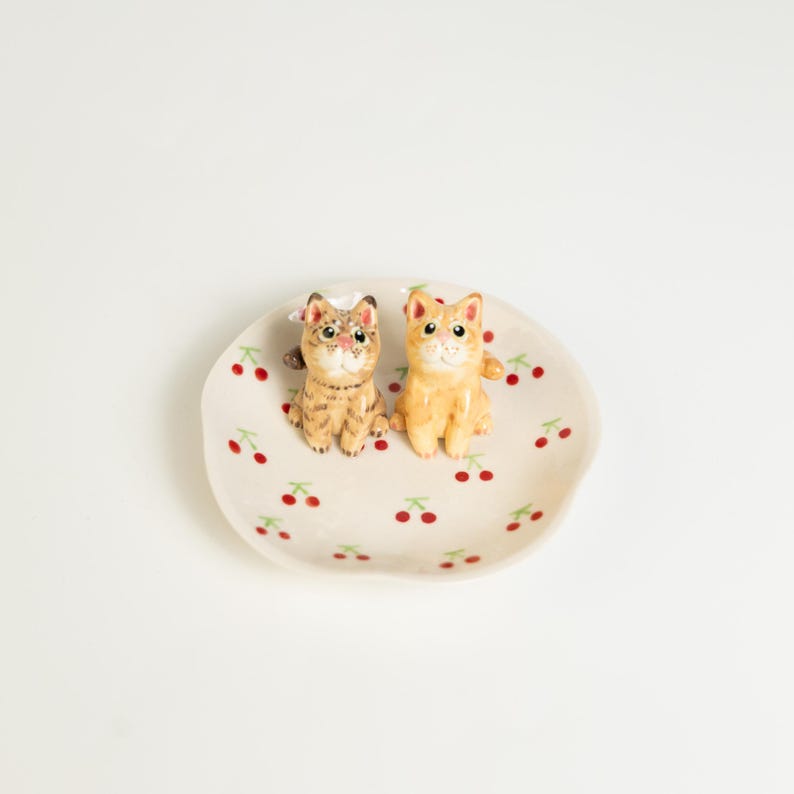 May include: A small, off-white ceramic dish with a scalloped edge, decorated with red cherries and green leaves. Two small cat figurines, one tan and one orange, sit on the dish. The cats have white bows on their heads.