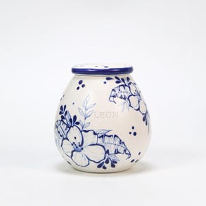 May include: A white ceramic jar with a lid, adorned with blue floral patterns and the name "LEON". The jar has a rounded shape and a dark blue rim. The floral design includes large flowers and leaves.