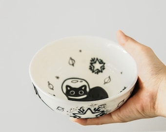 May include: White ceramic bowl with a black and white illustration of a cat wearing a space helmet and surrounded by stars, planets, and leaves.
