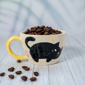May include: A yellow and white ceramic mug with a black cat design. The mug is filled with coffee beans.
