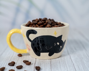 Puede incluir: Una taza de cerámica amarilla y blanca con un diseño de gato negro. La taza está llena de granos de café.