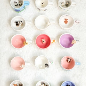 May include: A selection of ceramic mugs featuring dog figurines. The mugs are in various colours, including pink, blue, and white, and depict different dog breeds. Some mugs have dog figurines on the handles. The mugs are displayed on a white surface.