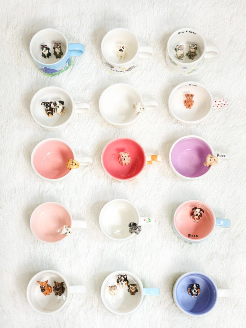 May include: A selection of ceramic mugs featuring dog figurines. The mugs are in various colours, including pink, blue, and white, and depict different dog breeds. Some mugs have dog figurines on the handles. The mugs are displayed on a white surface.