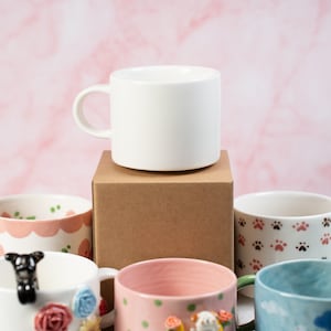 Può includere: Una collezione di cinque tazze in ceramica con diversi design. Le tazze sono disposte su una scatola di cartone marrone. Una tazza è bianca e vuota, mentre le altre presentano motivi e design colorati, tra cui fiori, impronte di zampe e un cane dei cartoni animati.