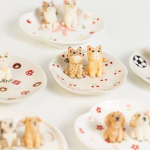 May include: A selection of small ceramic dishes, each adorned with a different animal figurine. The white dishes feature colourful patterns such as cherries, flowers, and a football. The figurines include cats and dogs.