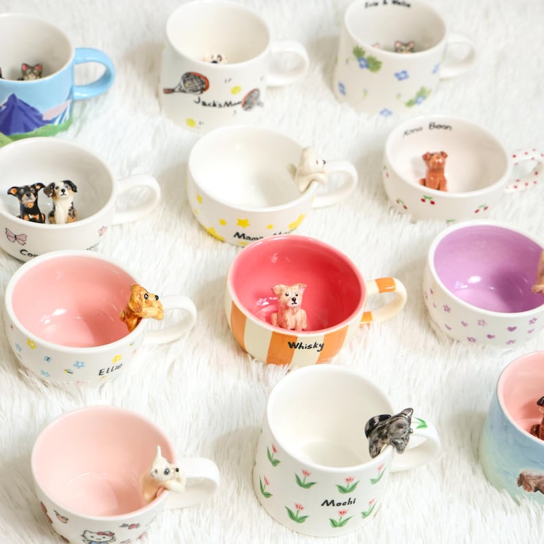 May include: A selection of ceramic mugs, each adorned with a dog figurine. The mugs come in various colours, including pink and blue, and feature different patterns like flowers and stars. Some mugs have text. The mugs are ideal for tea or coffee.