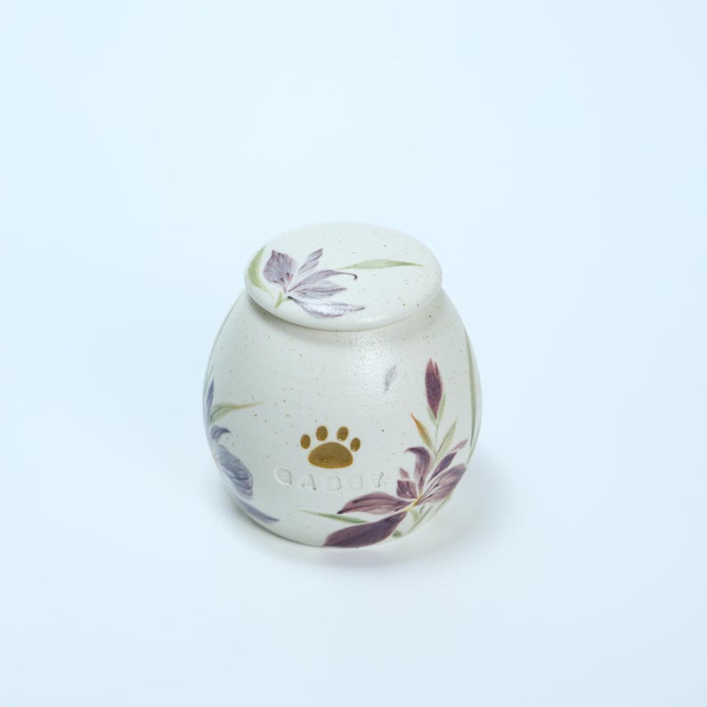 May include: White ceramic pet urn with a floral design and a gold paw print. The urn is inscribed with the name "Gaddy".