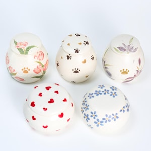 Peut inclure: Cinq pots en céramique avec couvercles, chacun décoré d'un motif différent. Les motifs comprennent des cœurs, des fleurs, des empreintes de pattes et un motif floral. Les pots sont blancs avec un glaçage crème.