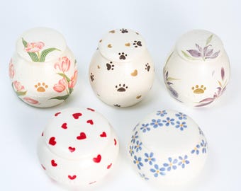 May include: Five ceramic jars with lids, each decorated with a different pattern. The patterns include hearts, flowers, paw prints, and a floral design. The jars are white with a cream-colored glaze.