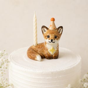 May include: A ceramic fox figurine cake topper featuring a brown and white fox wearing a tan party hat with orange dots and a yellow star pendant necklace. A thin, cream-colored spiral candle is inserted into a holder on the fox's back.