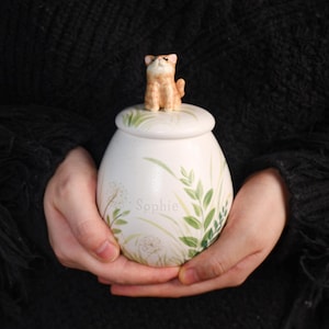 May include: A white ceramic urn with a lid, featuring a small, tan cat figurine on top. The urn is decorated with green leaf and floral designs, and the name "Sophie" is written on the side.