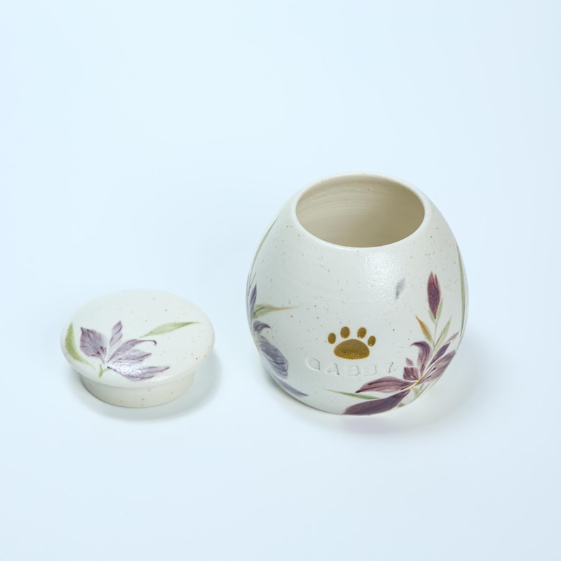 May include: White ceramic pet urn with a floral design featuring purple and green flowers. The urn has a lid and a gold paw print with the word "Gabby" inscribed on the front.