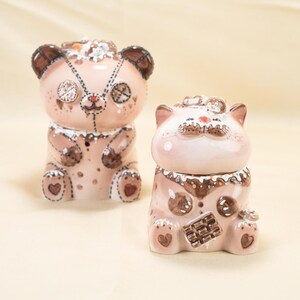 May include: Two ceramic bear and cat figurines. The bear is light pink with brown accents, decorative stitching, and heart-shaped feet. The cat is light pink with chocolate-colored accents and a chocolate bar design. Both have decorative embellishments.