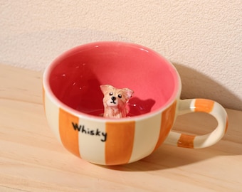 Puede incluir: Una taza de cerámica con una figura de perro en su interior. La taza tiene rayas verticales naranjas y crema, un interior rosa y un asa a juego. La palabra "Whisky" está escrita en el lateral.