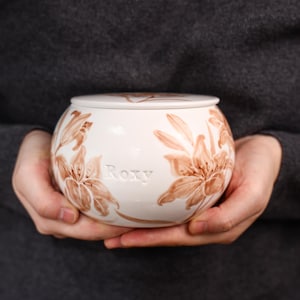 May include: A white ceramic urn with a lid, featuring brown floral designs and the name "Roxy". The urn is round and held in hands, with a dark gray background.