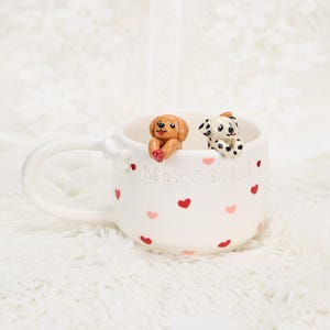 May include: White ceramic mug with a handle, decorated with red and pink hearts. Two small dog figurines, one brown and one Dalmatian, are perched on the rim. The mug has the text "Max & Mini".