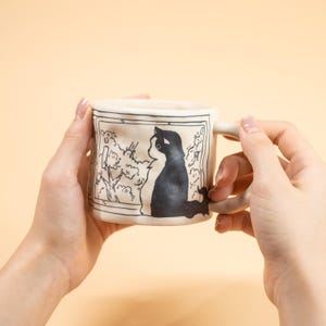 Peut inclure: Une tasse en céramique avec une illustration de chat noir et blanc. La tasse présente un design rectangulaire avec un contour noir et une anse. Le chat est représenté de profil, regardant une scène d'arbres et de nuages.