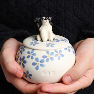 May include: A ceramic jar with a lid, adorned with a sitting dog figurine. The jar is white with blue floral patterns and a small text. The dog is black and white. The jar is held in two hands.