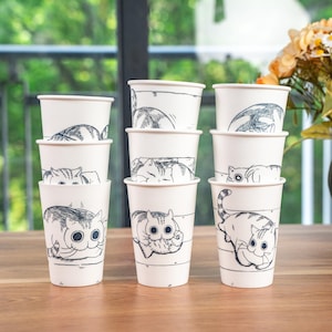 Peut inclure: Ensemble de huit gobelets en papier blanc avec des dessins au trait noir de chats. Les chats sont dans différentes poses, notamment en train de dormir, de s'asseoir et de jouer.