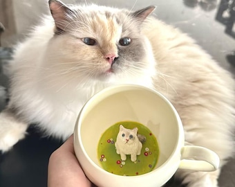 May include: A white ceramic mug with a green and yellow floral design inside. A small white cat figurine is sitting in the center of the mug.