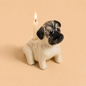以下が含まれることがあります： クリーム色の体にグレーの陰影と黒い顔を持つ、陶器製のパグの置物型キャンドルホルダー。パグは座った姿勢をしており、背中の穴には小さな白いテーパーキャンドルが立てられ、火が灯されています。実用的なインテリアアイテムです。