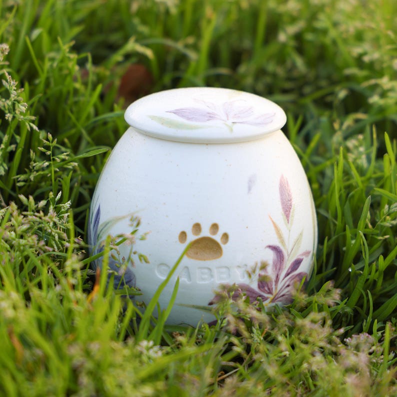 May include: White ceramic pet urn with a floral design and a paw print. The urn has a lid and the name "Abby" is inscribed on the front.