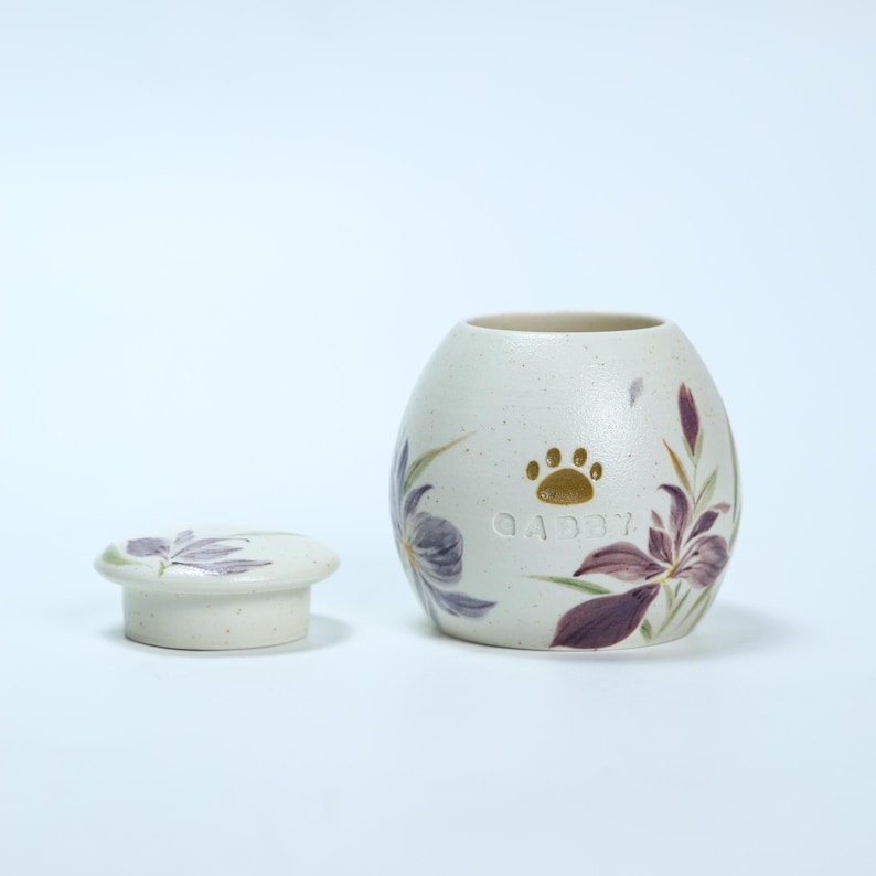 May include: White ceramic pet urn with a floral design in shades of purple and green. The urn has a gold paw print and the name "Gabby" inscribed on it. The urn comes with a matching lid.
