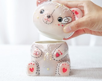 May include: A pink ceramic bear-shaped container with a removable lid. The bear has black stitched details, heart accents, and a decorative design. The lid is being lifted to reveal the inside of the container.