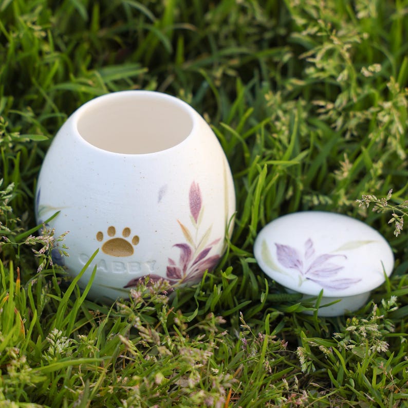 May include: White ceramic pet urn with a floral design and a paw print. The urn has the name "Abby" inscribed on it. The lid is off and sitting next to the urn.